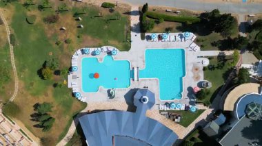 Güneşli bir günde çekilmiş şezlonglar, şemsiyeler ve küçük bir su parkı olan açık havuz kompleksinin yukarıdan aşağı havuzu görüntüsü..