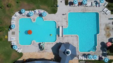 Güneşli bir günde çekilmiş şezlonglar, şemsiyeler ve küçük bir su parkı olan açık havuz kompleksinin yukarıdan aşağı havuzu görüntüsü..