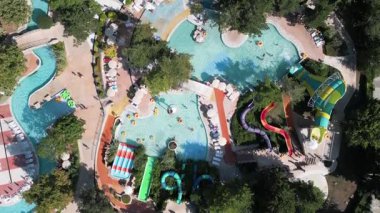 Güneşli bir günde yemyeşil ve dinlenme yerleriyle çevrili dolambaçlı slaytlar, havuzlar ve tembel nehirlerle dolu renkli bir su parkının hava aracı görüntüsü..