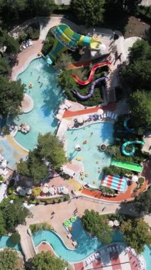 Güneşli bir günde yemyeşil ve dinlenme yerleriyle çevrili dolambaçlı slaytlar, havuzlar ve tembel nehirlerle dolu renkli bir su parkının hava aracı görüntüsü..