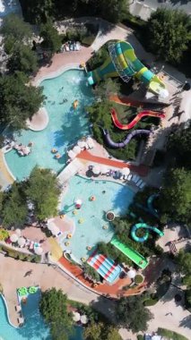 Güneşli bir günde yemyeşil ve dinlenme yerleriyle çevrili dolambaçlı slaytlar, havuzlar ve tembel nehirlerle dolu renkli bir su parkının hava aracı görüntüsü..
