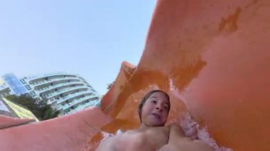 Güneşli bir günde parlak kırmızı bir su kaydırağında kayan mutlu bir çocuğun video görüntüsü. Aquapark 'ta yaz eğlencesinin tadını çıkarıyor ve sıçrıyor..