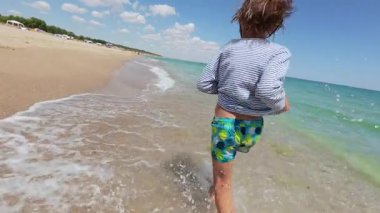 Slow motion video of a child running along the beach shoreline on a sunny summer day. Waves splash around the feet. Joyful and carefree moment of childhood vacation by the sea.