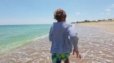Slow motion video of a child running along the beach shoreline on a sunny summer day. Waves splash around the feet. Joyful and carefree moment of childhood vacation by the sea.