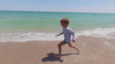 Slow motion video of a happy child running barefoot along a sandy beach on a sunny summer day. Joyful summer vibes, carefree childhood, coastal vacation atmosphere.