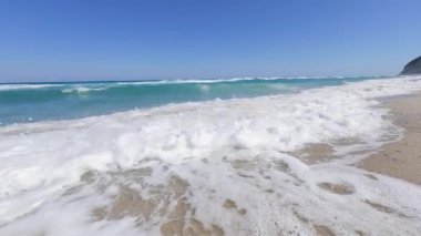 Slow motion footage of a powerful sea wave breaking near the camera under a clear blue sky. Crystal clear turquoise water and foamy splash in bright daylight.