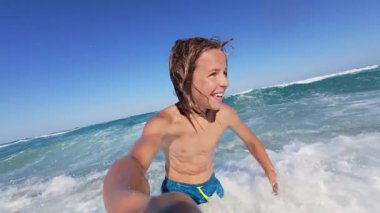 Slow motion video of a happy boy splashing water in the sea, smiling at the camera on a bright sunny day. Pure joy and summer fun at the beach, captured in vivid detail.