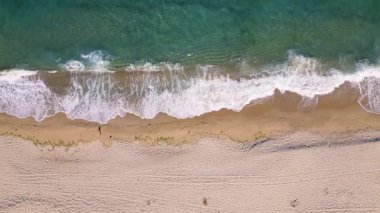 Aerial view of ocean waves gently rolling onto an empty sandy beach. Clear turquoise water, white foam, and soft golden sand create a peaceful coastal scene. Ideal for travel or nature themes.