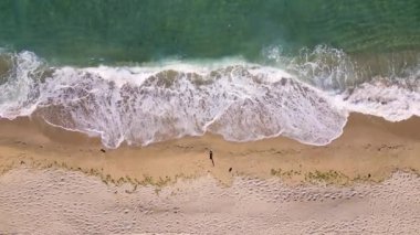 Aerial view of ocean waves gently rolling onto an empty sandy beach. Clear turquoise water, white foam, and soft golden sand create a peaceful coastal scene. Ideal for travel or nature themes.