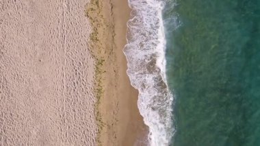 Aerial view of ocean waves gently rolling onto an empty sandy beach. Clear turquoise water, white foam, and soft golden sand create a peaceful coastal scene. Ideal for travel or nature themes.