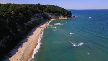 Aerial footage of a scenic sandy beach with waves gently hitting the shore, people relaxing under umbrellas, and green cliffs in the background. Peaceful summer vibe captured by drone.