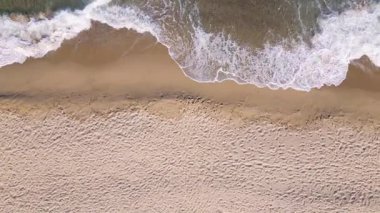 Aerial view of ocean waves gently rolling onto an empty sandy beach. Clear turquoise water, white foam, and soft golden sand create a peaceful coastal scene. Ideal for travel or nature themes.