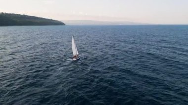 Aerial video of a white sailboat sailing alone on deep blue sea. Calm waves shimmer under sunlight. Peaceful and adventurous maritime scene perfect for travel and freedom themes.