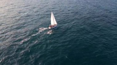 Aerial video of a white sailboat sailing alone on deep blue sea. Calm waves shimmer under sunlight. Peaceful and adventurous maritime scene perfect for travel and freedom themes.