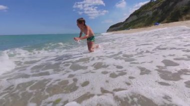 Slow motion video of happy child playing in shallow sea waves, smiling and enjoying summer day at beach with clear water and bright sky.