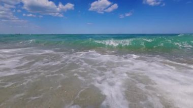 Slow motion POV shot of powerful ocean wave crashing near shore, white foam and clear turquoise water under blue sky with scattered clouds.
