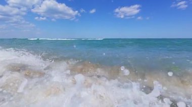 Slow motion POV shot of powerful ocean wave crashing near shore, white foam and clear turquoise water under blue sky with scattered clouds.