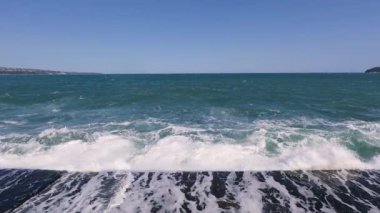 Slow motion video of a powerful ocean wave crashing against a pier, creating dramatic white spray under a clear blue sky.