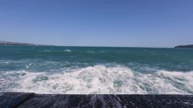 Slow motion video of a powerful ocean wave crashing against a pier, creating dramatic white spray under a clear blue sky.