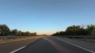 Driving on a highway at sunset with clear blue sky, trees, and guardrails. POV car perspective, summer road trip mood.