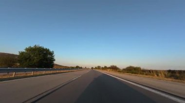 Driving on a highway at sunset with clear blue sky, trees, and guardrails. POV car perspective, summer road trip mood.