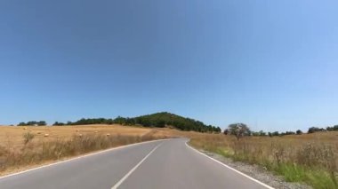 Driving along empty country road towards a green hill, surrounded by golden hay fields under clear blue sky. Scenic rural travel video.