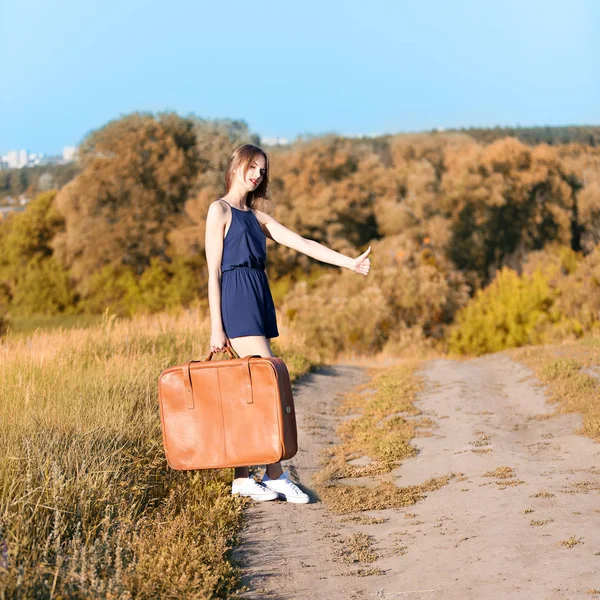 Genç güzel kız bir ülke yolunda ve oy kenarda el ile bir yükseltilmiş ile ayakta, tonlama bir bavulla ince.