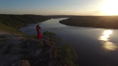 Hava uçuş uzak çift cliff Edge suya yakın duran aşık. Şaşırtıcı kırmızı elbiseli kadın. Siyah adam. Günbatımı.