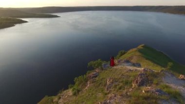 Uçurum kenarı suya yakın ayakta aşık çift yukarıda hava uçuş. Şaşırtıcı kırmızı elbiseli kadın. Siyah adam. Günbatımı.