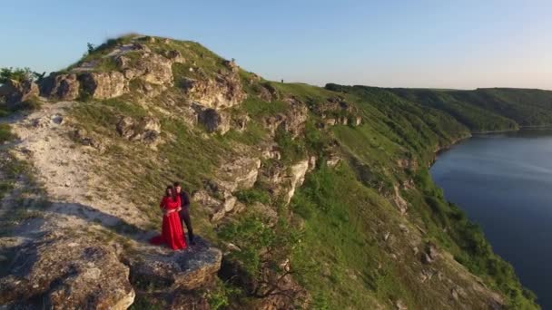 Aerial flight away from couple in love standing in cliff edge near water. Woman in amazing red dress. Man in black. Sunset. — Stock Video