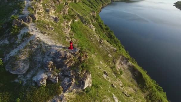 Vuelo aéreo alrededor de pareja enamorada de pie en el borde del acantilado cerca del agua. Mujer con un vestido rojo increíble. Hombre de negro. Puesta de sol . — Vídeos de Stock