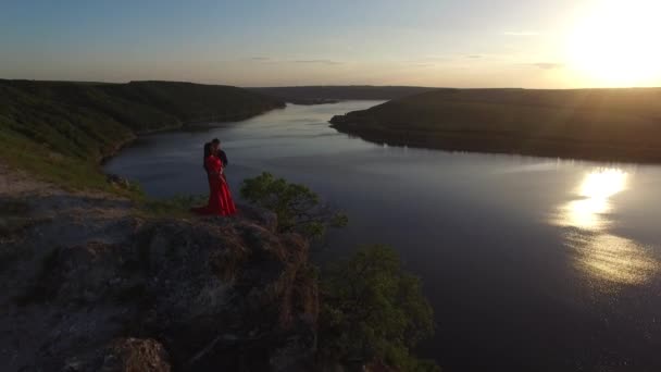 Vuelo aéreo lejos de la pareja enamorada de pie en el borde del acantilado cerca del agua. Mujer con un vestido rojo increíble. Hombre de negro. Puesta de sol . — Vídeos de Stock