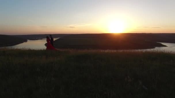 Letecké letu poblíž pár v lásce chůze na okraji řeky banky. Žena v úžasných červených šatech. Muž v černém. Západ slunce. — Stock video