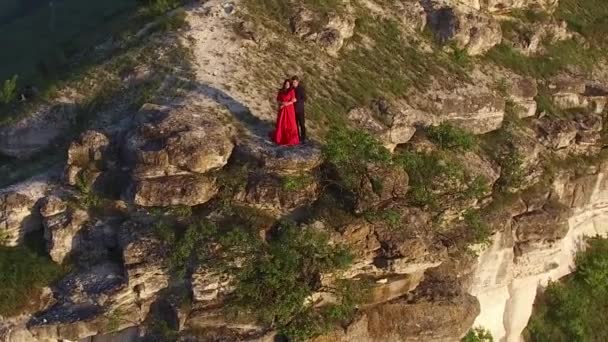 Luchtfoto vlucht rond paar verliefd permanent in de rand van de klif in de buurt van water. Vrouw in geweldige rode jurk. Man in het zwart. Zonsondergang. — Stockvideo