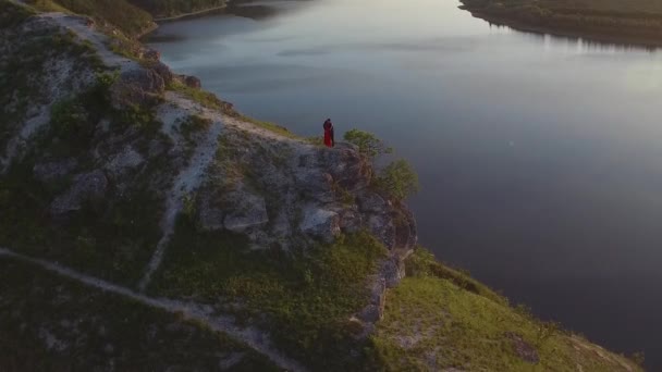 Luchtfoto vlucht boven paar verliefd zoenen in de rand van de klif in de buurt van water. Vrouw in geweldige rode jurk. Man in het zwart. Zonsondergang. — Stockvideo