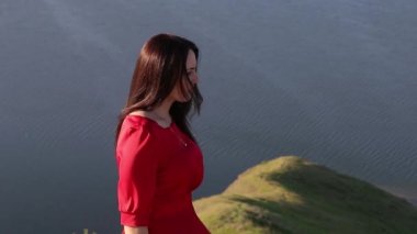 Nehir banka kenarına kamera için poz kırmızı elbiseli güzel beyaz kadın. Yaz gün batımı. Orta atış.