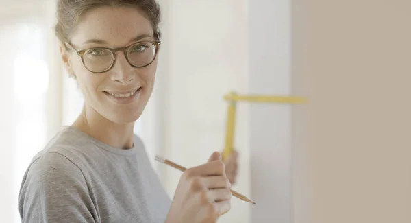 Smiling young woman doing a home makeover, she is measuring a wall using a folding ruler, DIY and house renovation concept