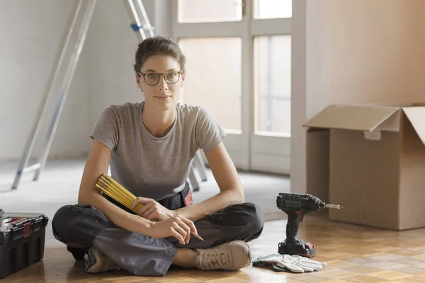Young woman moving in her new house and doing a home makeover, she is sitting on the floor with tools and cardboard boxes