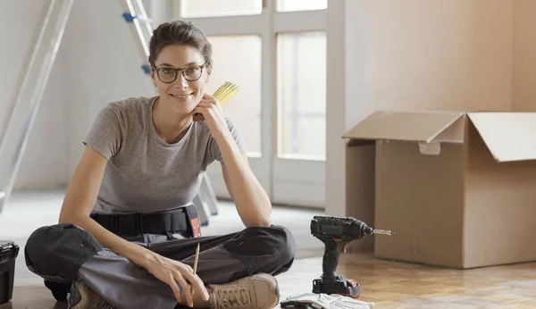 Young woman moving in her new house and doing a home makeover, she is sitting on the floor with tools and cardboard boxes