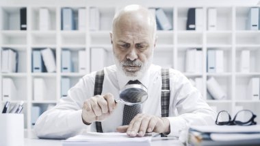 Senior corporate businessman reading paperwork using a magnifier, he is checking carefully an agreement  clipart