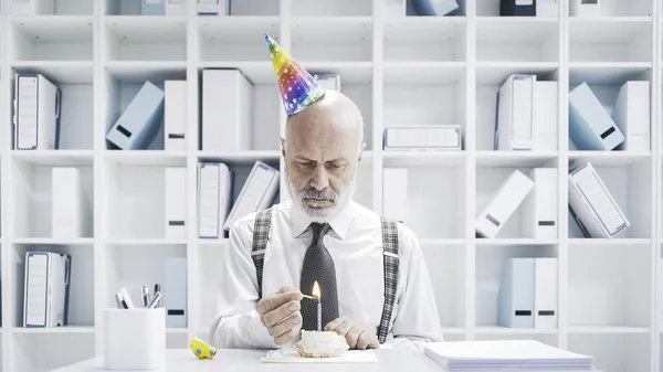 Senior businessman having a sad lonely birthday in the office, he is wearing a party hat and having a small cake alone