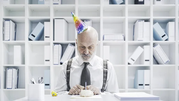 Senior businessman having a sad lonely birthday in the office, he is wearing a party hat and having a small cake alone