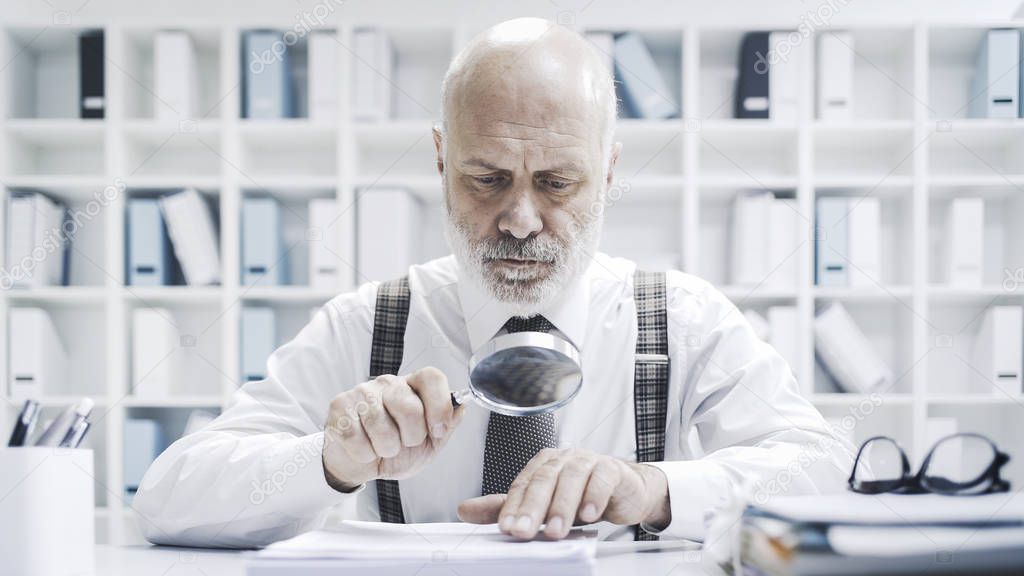 Senior corporate businessman reading paperwork using a magnifier, he is checking carefully an agreement 