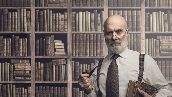 Confident senior academic professor smoking a pipe and posing in the library, old bookshelves in the background