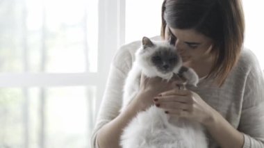 Bir pencere yanında oturan genç mutlu kadın o sevimli kedi onun kucağına holding ve karnı sürtünme