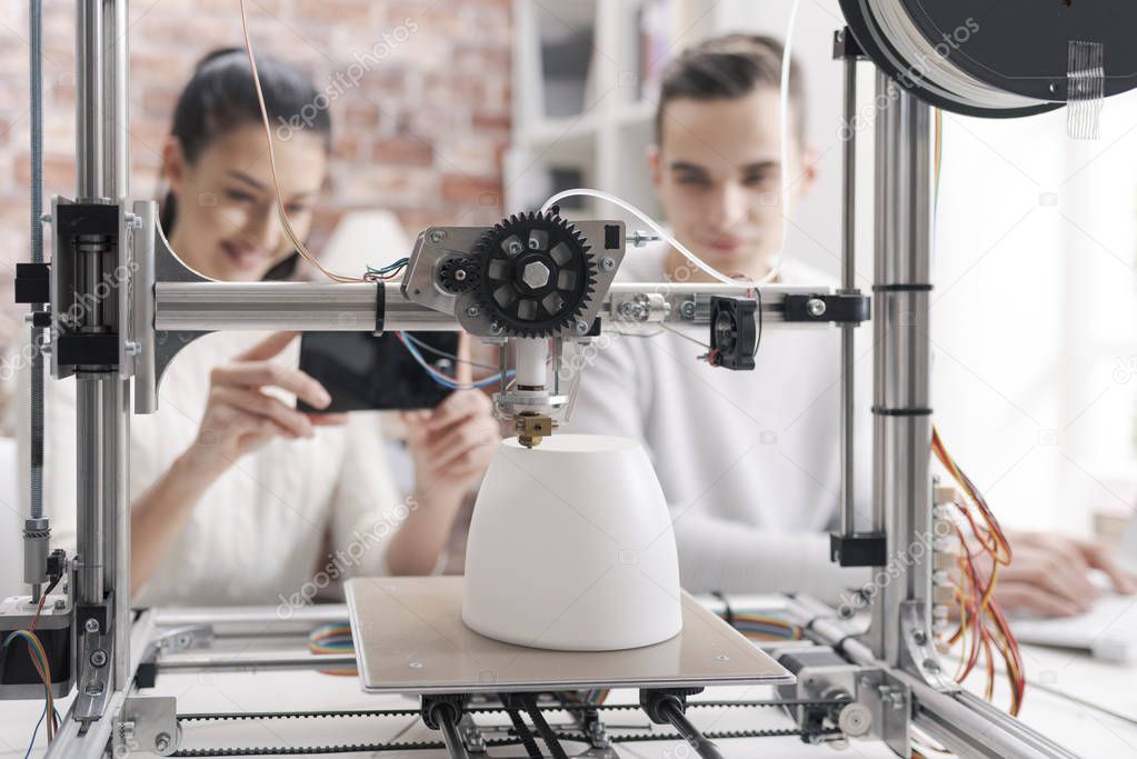 Engineering students printing prototype models using a 3D printer, the girl is taking pictures with a smartphone and sharing online