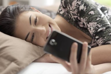 Genç gülümseyen kadın yatakta rahatlatıcı ve onun smartphone ile bağlanma, o arkadaşları ile sohbet ve iletileri okuma
