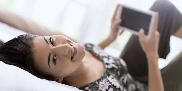 年轻快乐的女人在家中躺在床上放松 并与数字平板电脑联系 她正在网上观看视频 并在社交媒体上与朋友聊天 — 图库照片