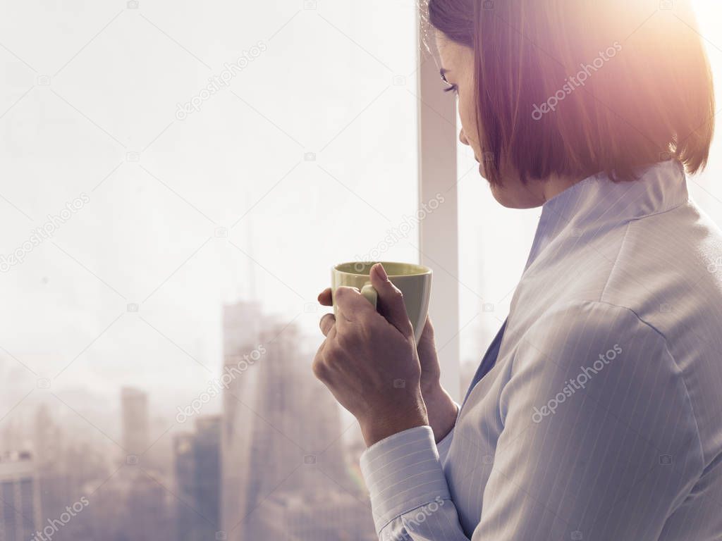 Businesswoman standing in front of a window and having a relaxing coffee break, she is holding a cup