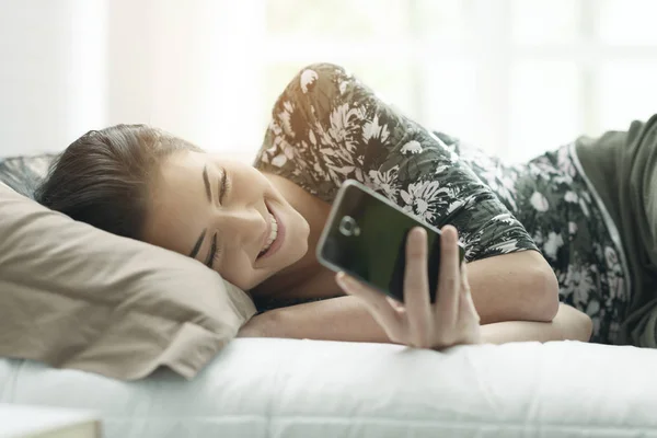 Молодая женщина лежит на кровати и соединяется со своим телефоном — стоковое фото